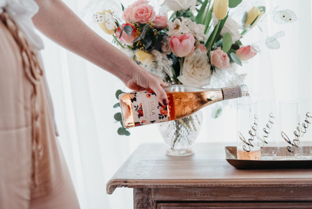 Women dressed in light blush pants pouring Pelee Island Winery Lola Cabernet Franc Rosé VQA Ontario into a glass that says cheers, sitting on a bar with a bouquet of flowers.