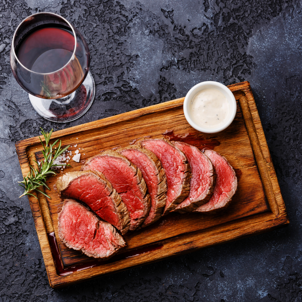 Wooden cutting board with sliced BBQ steak with rosemary sprig and a sauce with a glass of Pelee Island Winery Vinedressers Cabernet Sauvignon Petit Verdot VQA