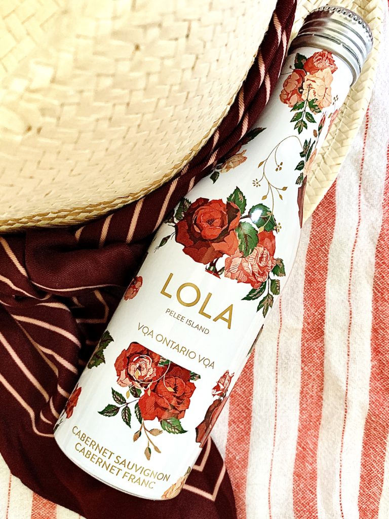 A 250mL aluminum bottle of LOLA Cabernet Sauvignon/Cabernet Franc blend sitting on top of a beach towel and sun hat.